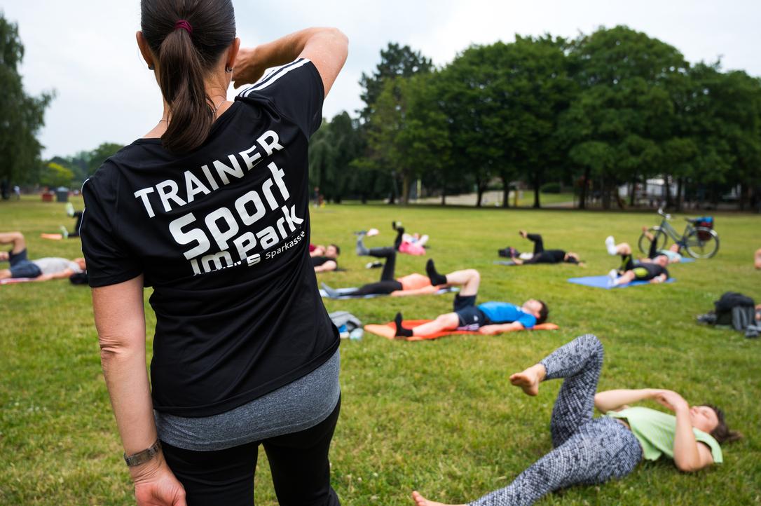 Trainerin mit schwarzem Shirt leitet Sportübungen im Park; Teilnehmer liegen auf Matten und trainieren im Hintergrund.