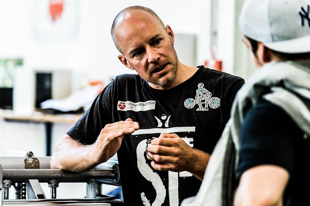 Mann mit glattem Kopf erklärt einem Gesprächspartner etwas in einem Sportstudio.