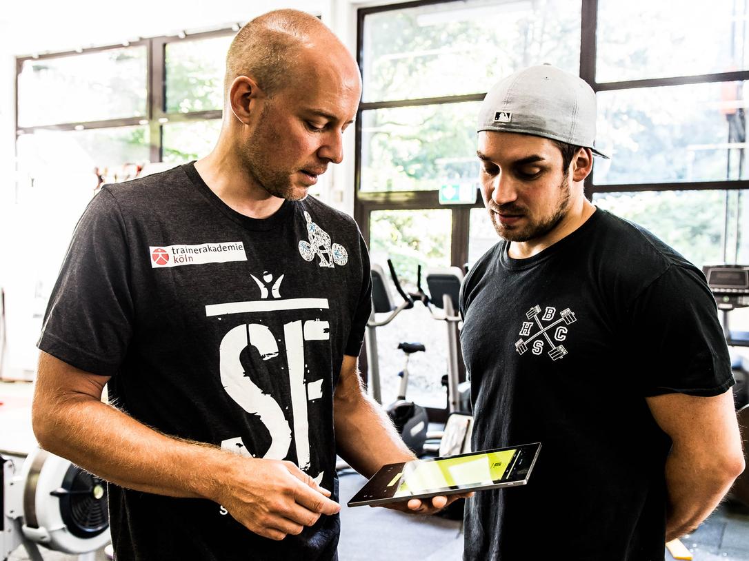 Zwei Trainer im Fitnessstudio betrachten gemeinsam ein Tablet. Im Hintergrund sind Trainingsgeräte zu sehen.