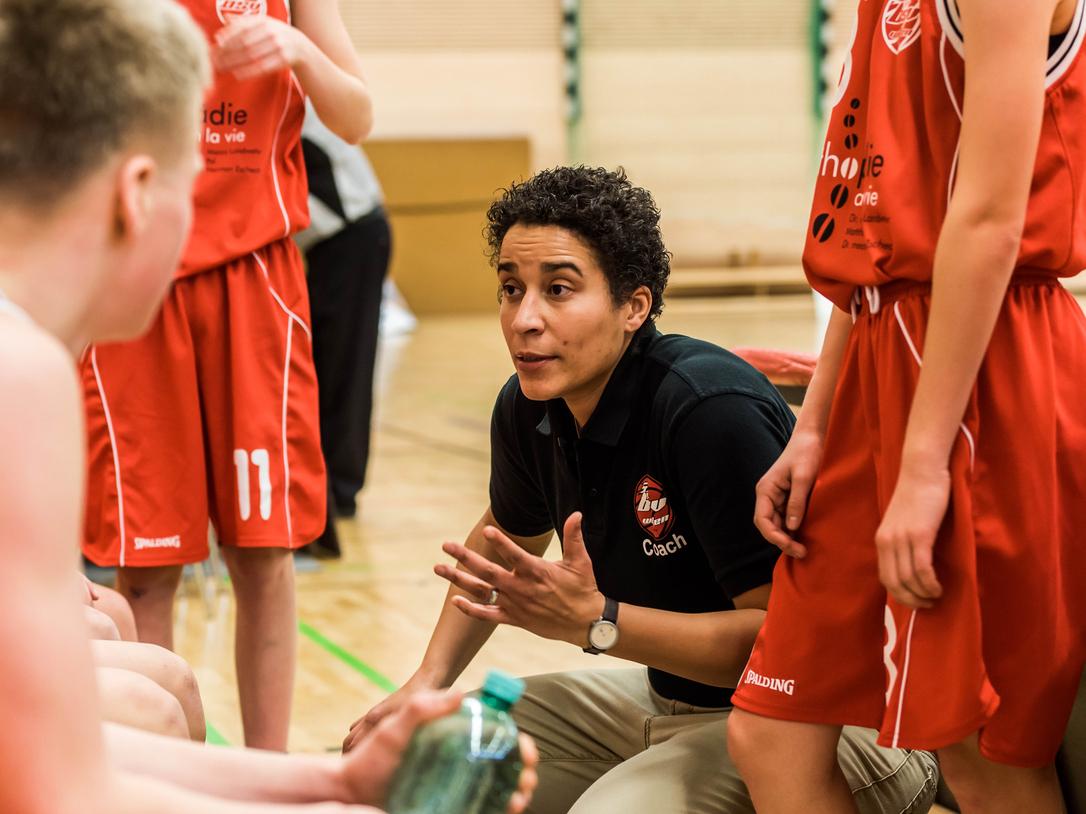 Basketballtrainerin erklärt Taktik während einer Auszeit zu Jugendlichen in roten Trikots auf dem Spielfeld.