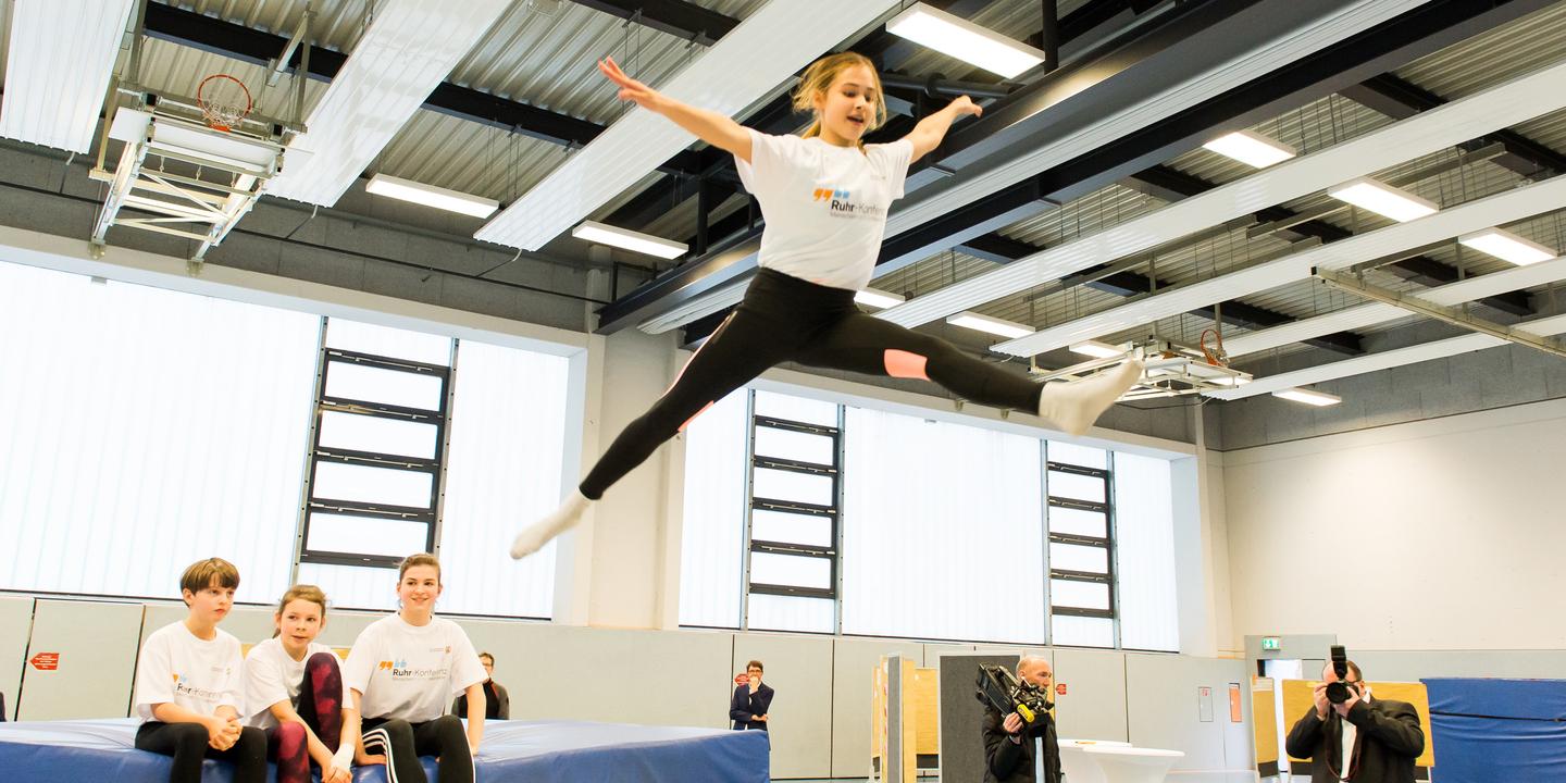 Mädchen springt mit ausgestreckten Armen über eine Trampolinfläche, während Zuschauer auf einer Bank sitzen.