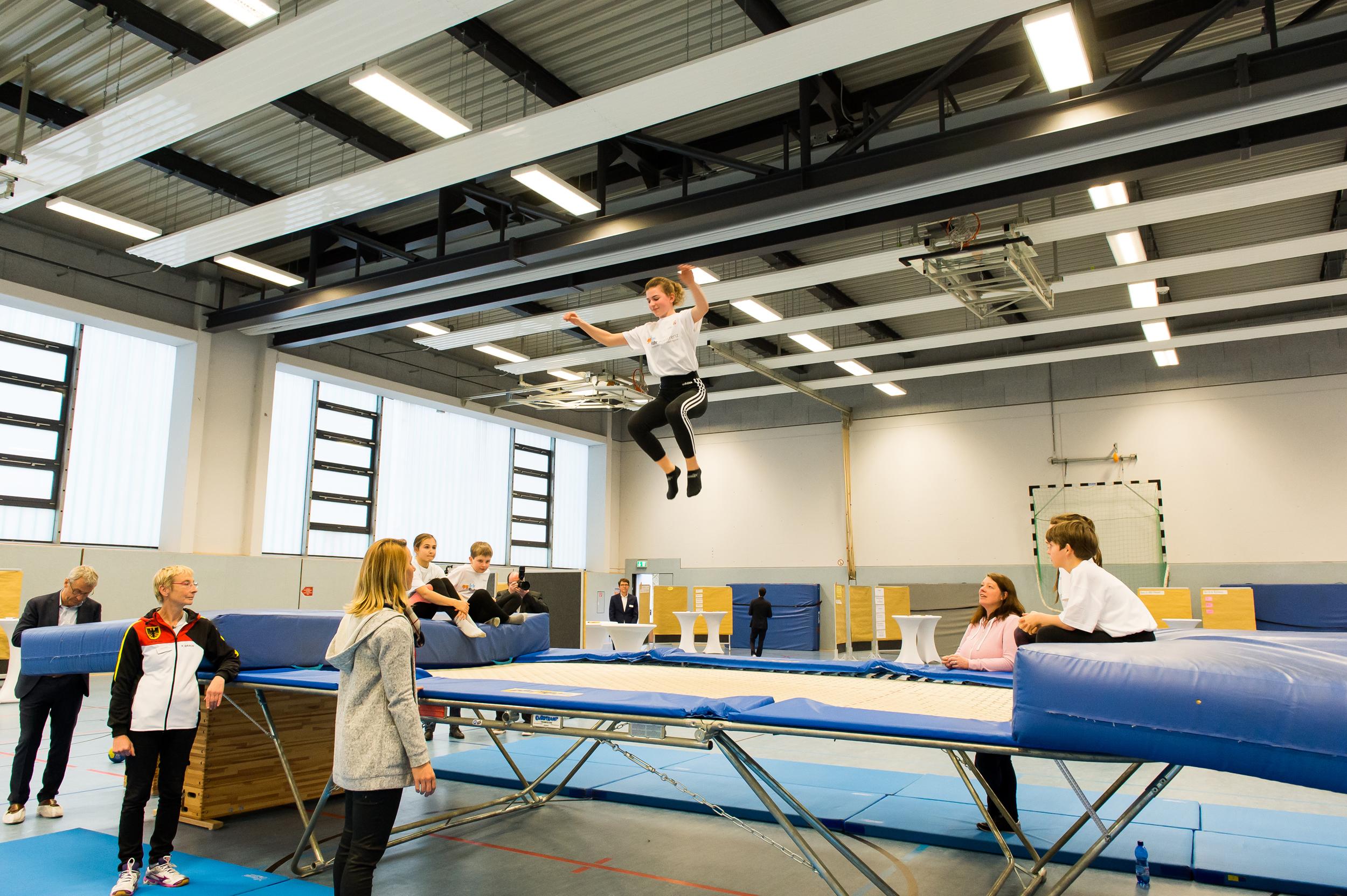 Junge springt auf einem Trampolin, während Zuschauer und Trainer im Hintergrund beobachten. Sporthalle mit hellem Licht.