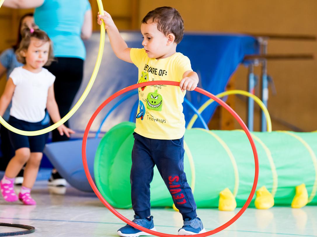 Zwei Kinder spielen in einer Turnhalle mit hula-hoops und einem grünen Tunnel. Ein Kind hält zwei Reifen in der Hand.