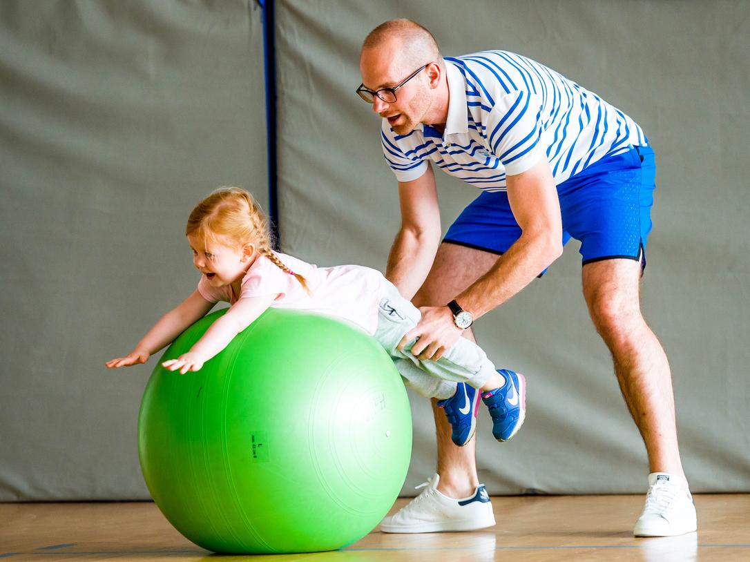 Ein Erwachsener hilft einem Kind, auf einem großen grünen Gymnastikball zu balancieren, in einer Sporthalle.