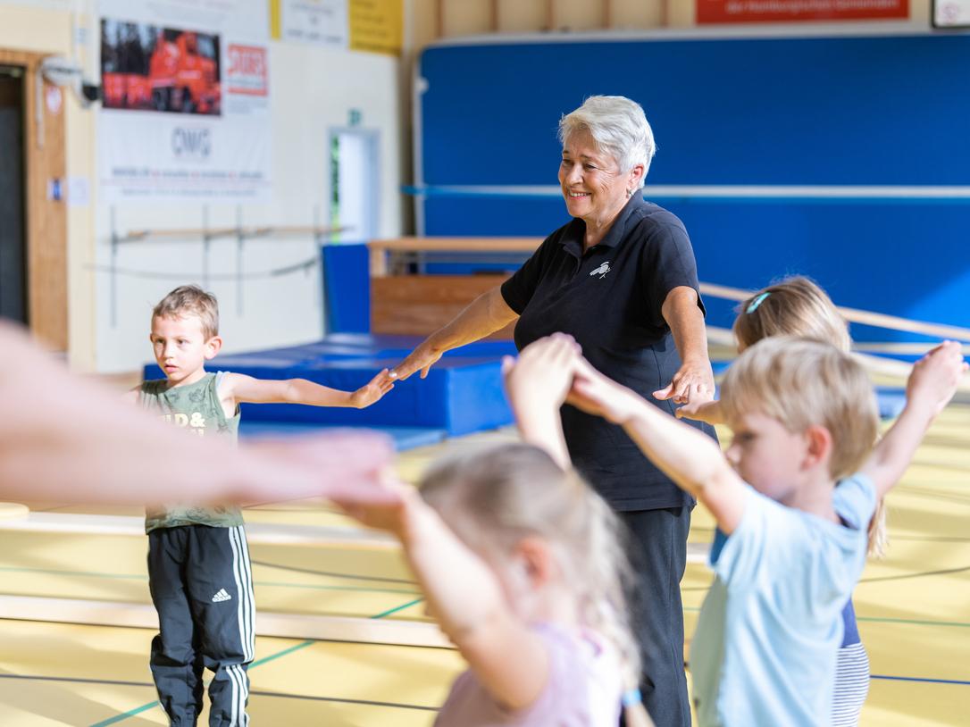 Seniorin leitet Kinder in einer Sporthalle an, während sie ihre Arme in die Höhe strecken.